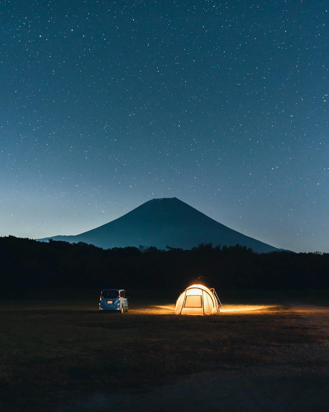 Carpa iluminada y microbús en pradera; el Monte Fuji en silueta bajo cielo estrellado en noche despejada