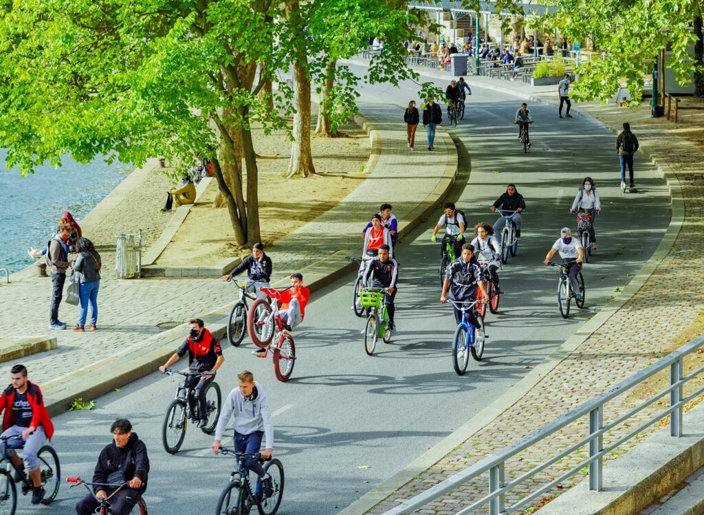 一群人沿著河畔的自行車道騎行，有男有女，年齡各異，旁邊是綠樹成蔭的人行道，行人正在散步